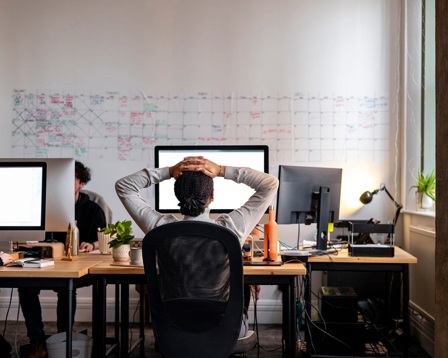 View from behind of a woman sitting at an office desk with her hands on her head